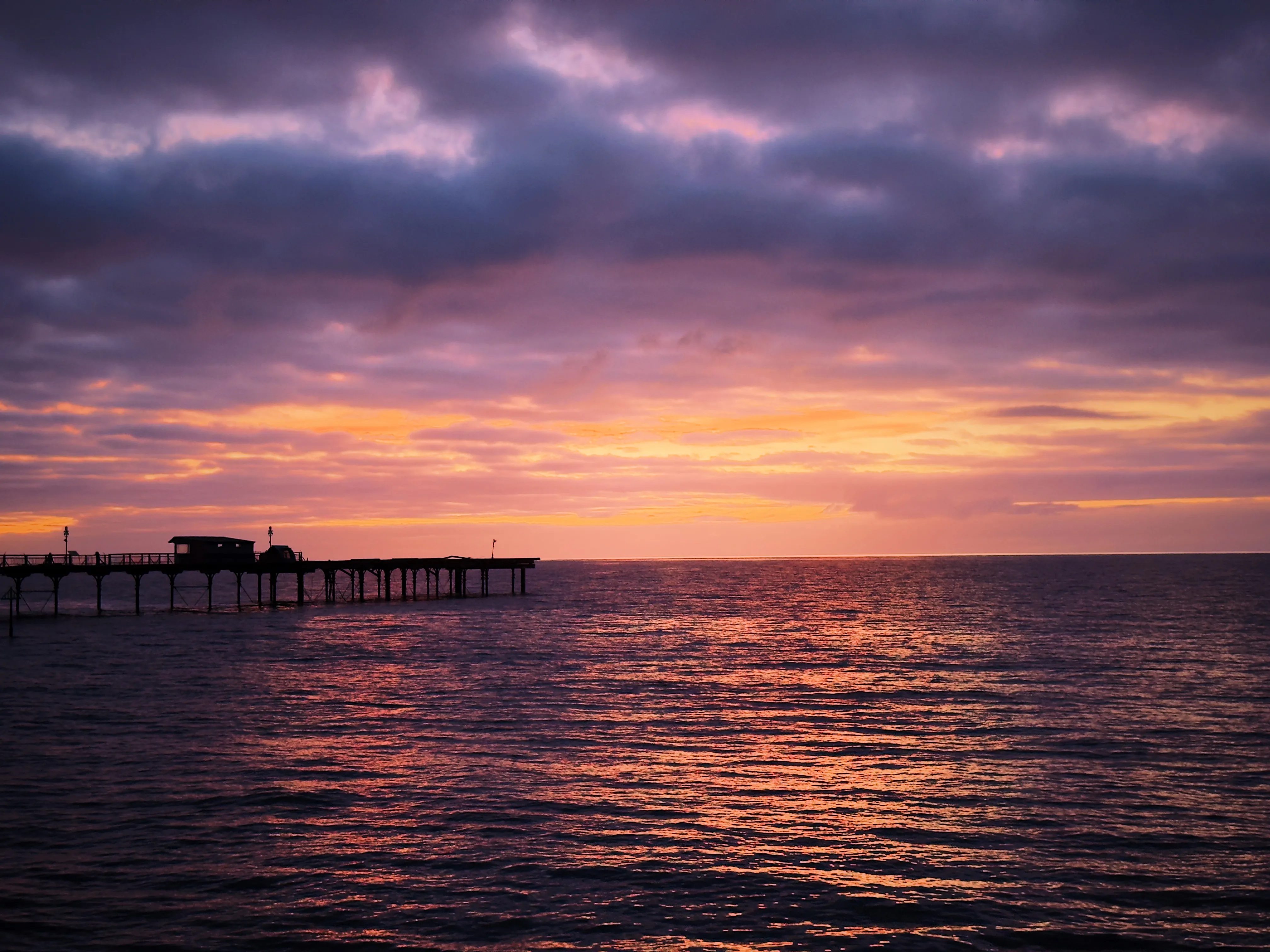 Vivid post-sunrise sky in deep purple, magenta and orange, a Victorian pier silhouetted on the left, calm sea holding the colour