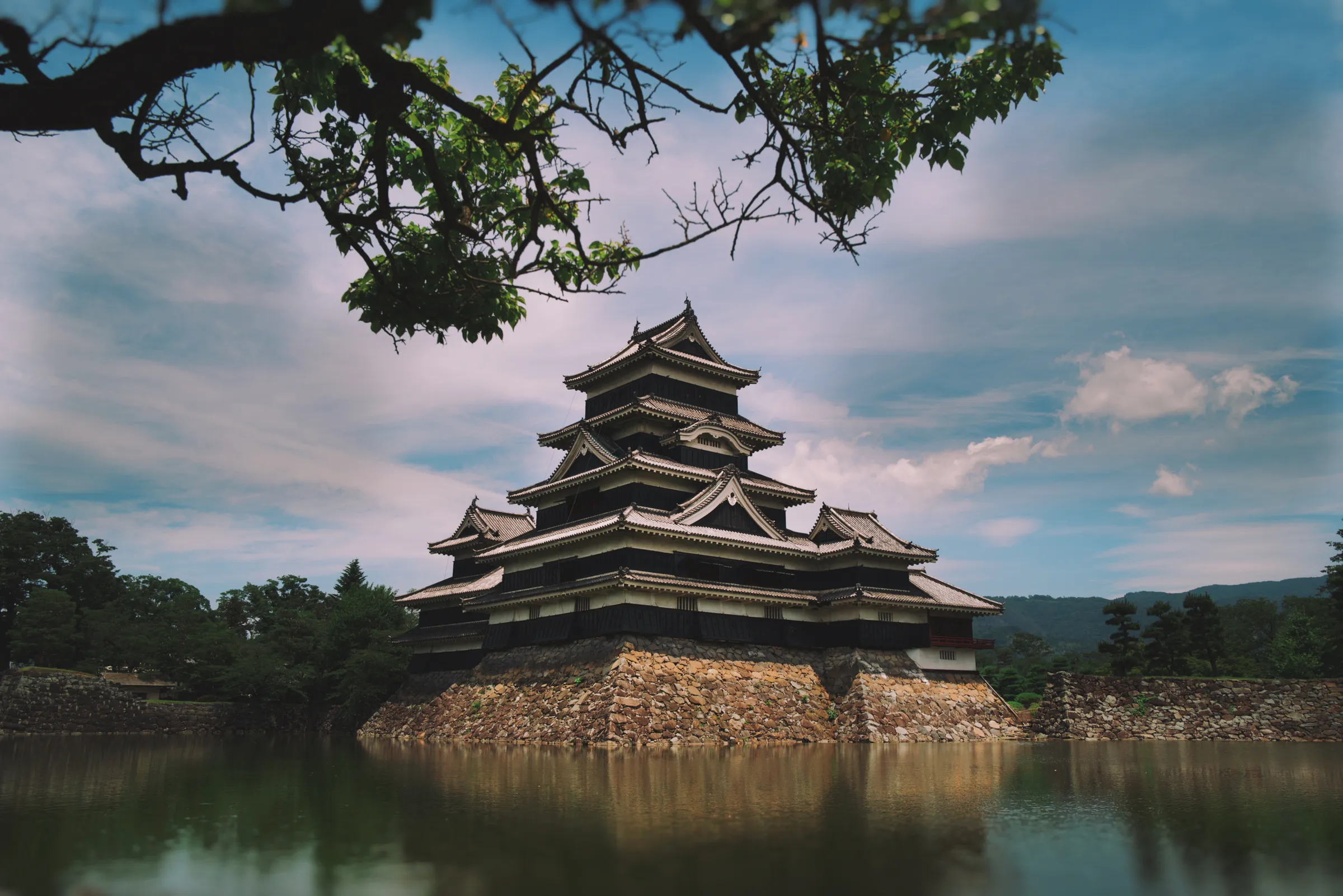 Matsumoto Castle rising from its moat, framed by overhanging branches, dramatic clouded sky