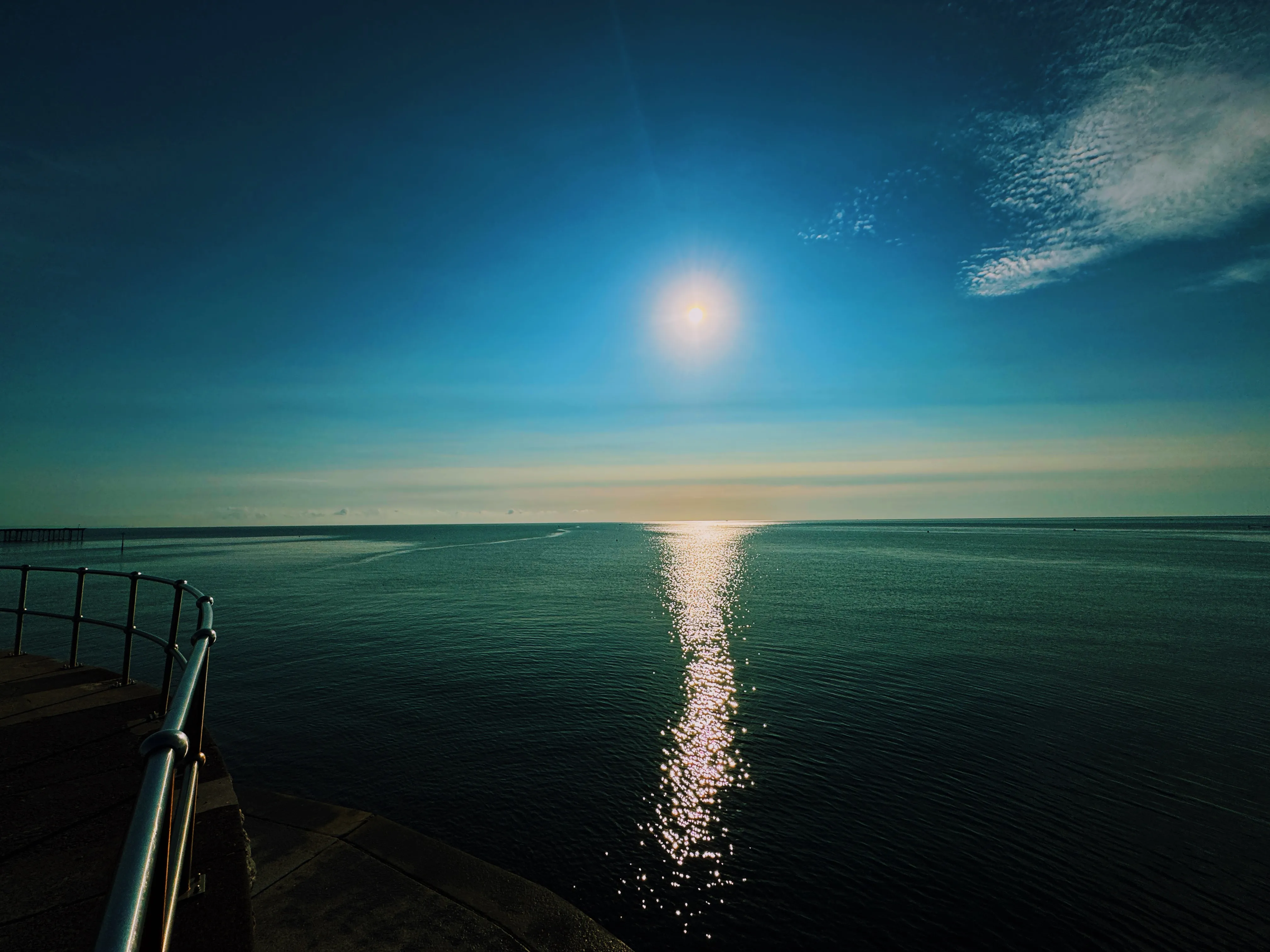 Sun lens flare at beach level in September, white foam churning in the foreground shallows, pale blue sky with thin cloud
