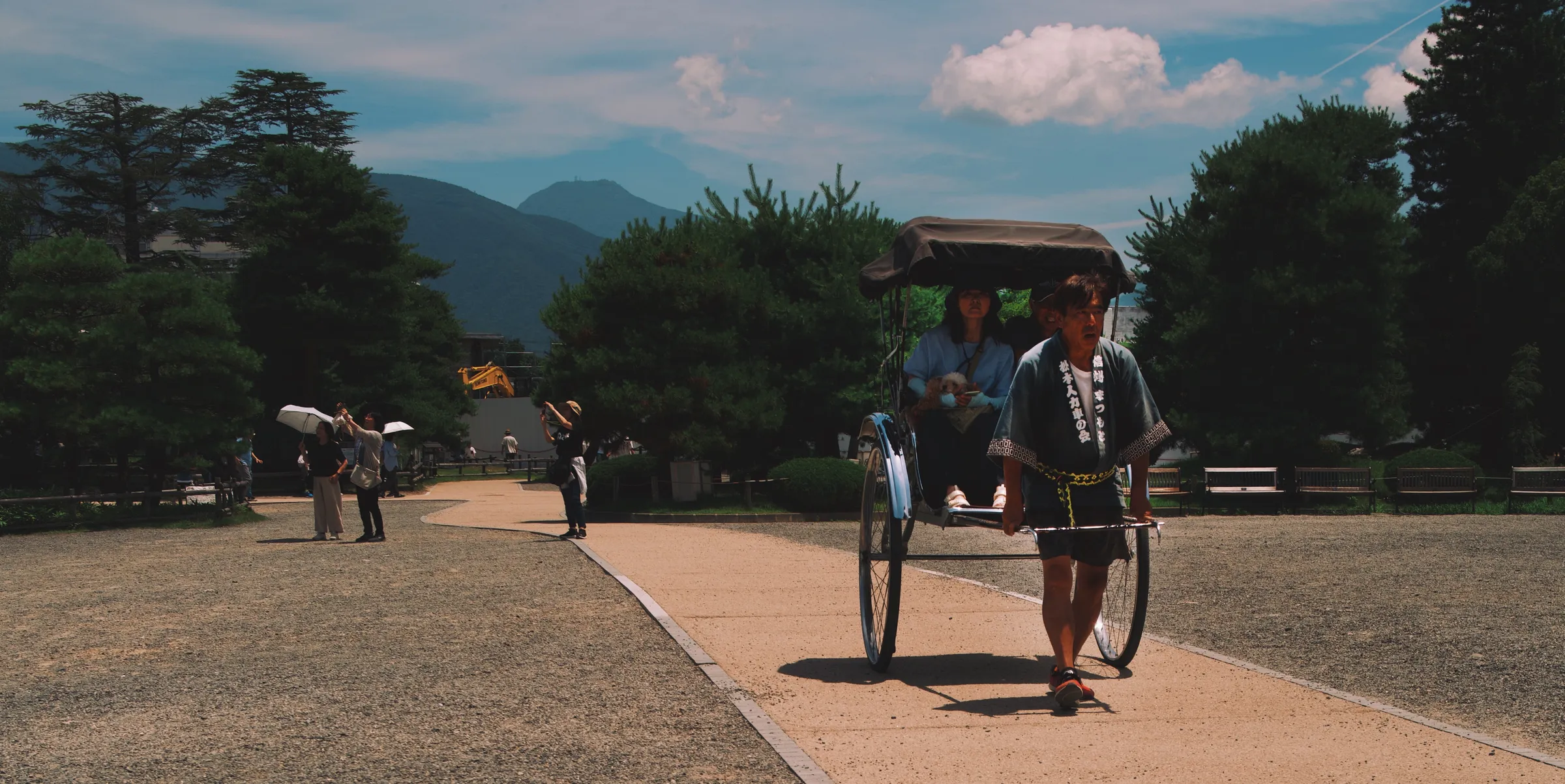 A rickshaw being pulled across open castle grounds, pine trees and mountains behind, blue sky
