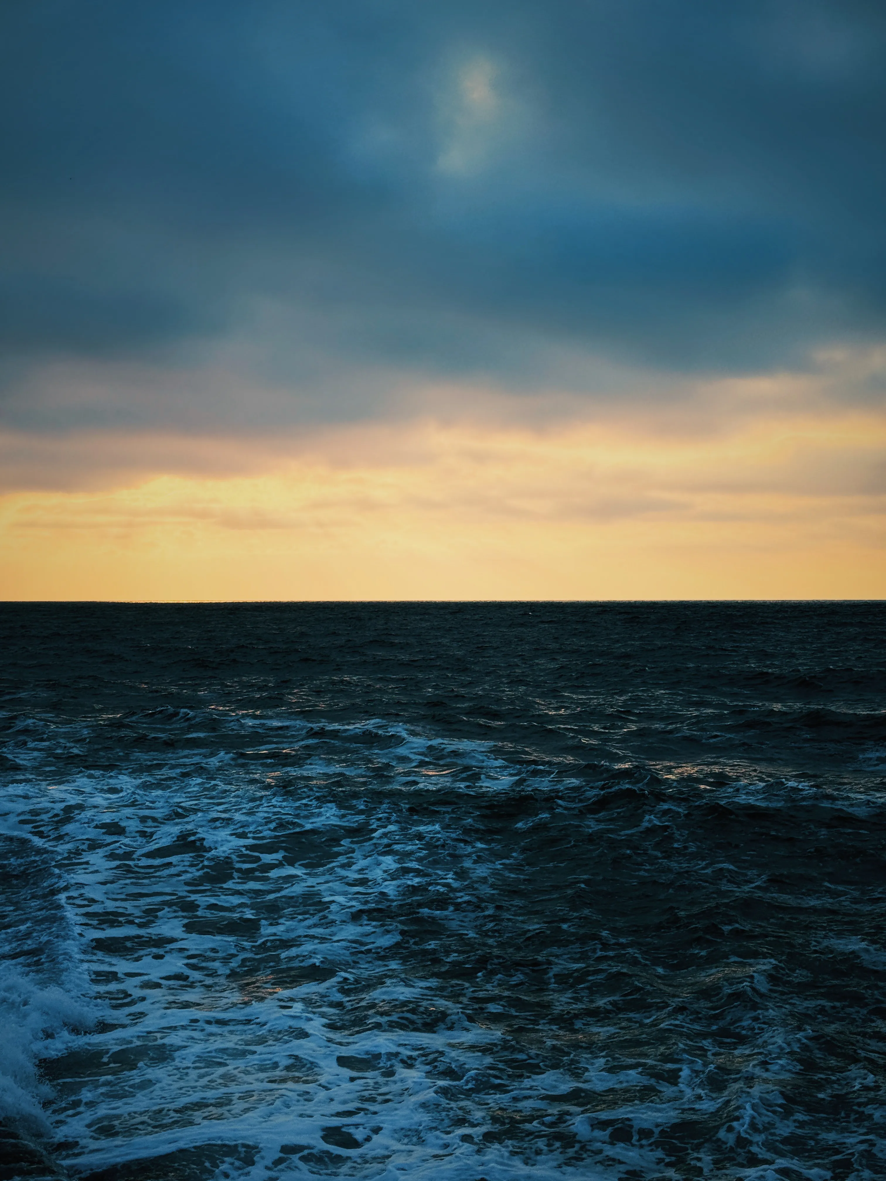 Dark stormy April dawn, heavy grey-blue cloud with a warm amber glow at the horizon, rough white-capped sea