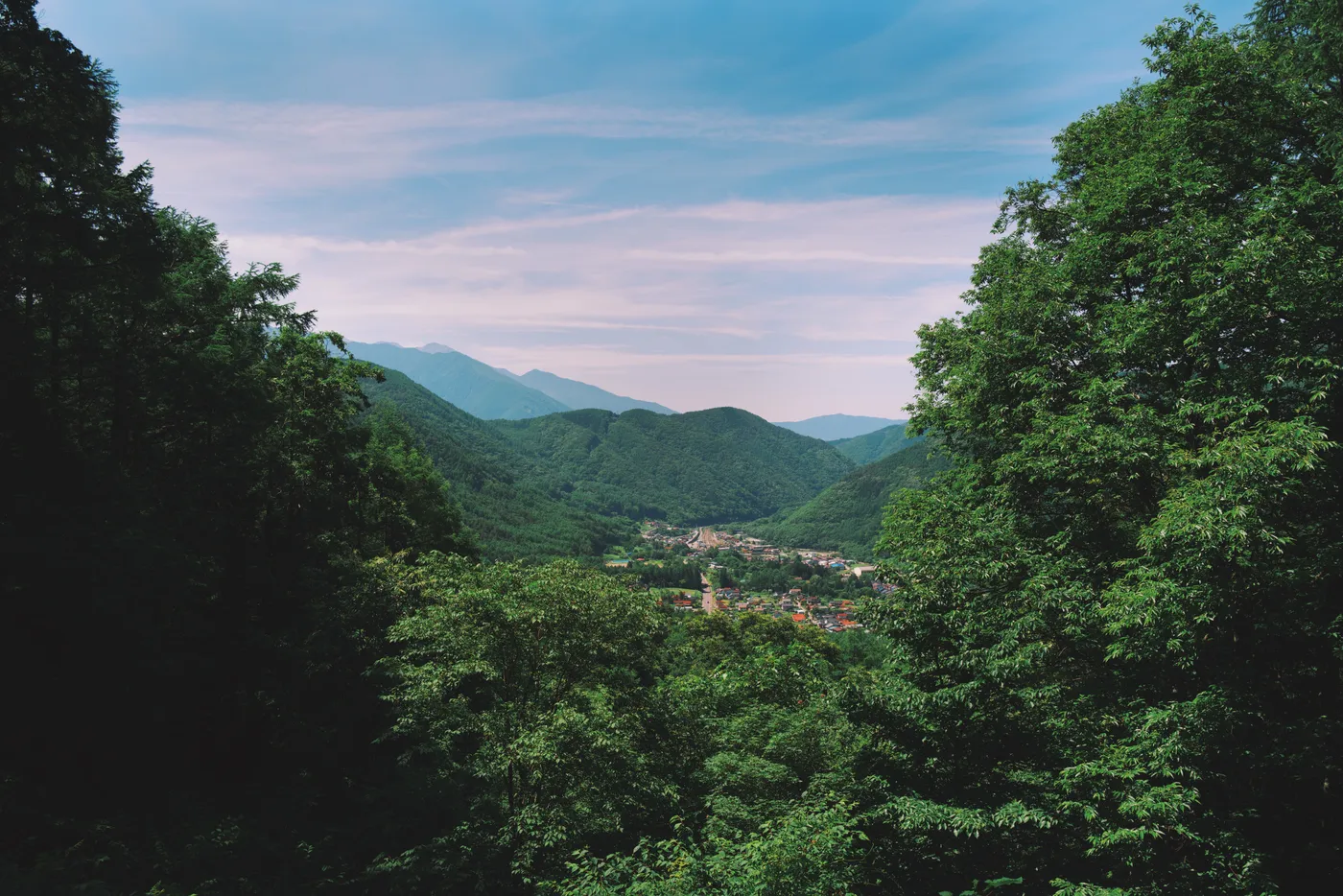Wide view across the Kiso Valley from a ridge, a settlement visible through the trees below, blue sky