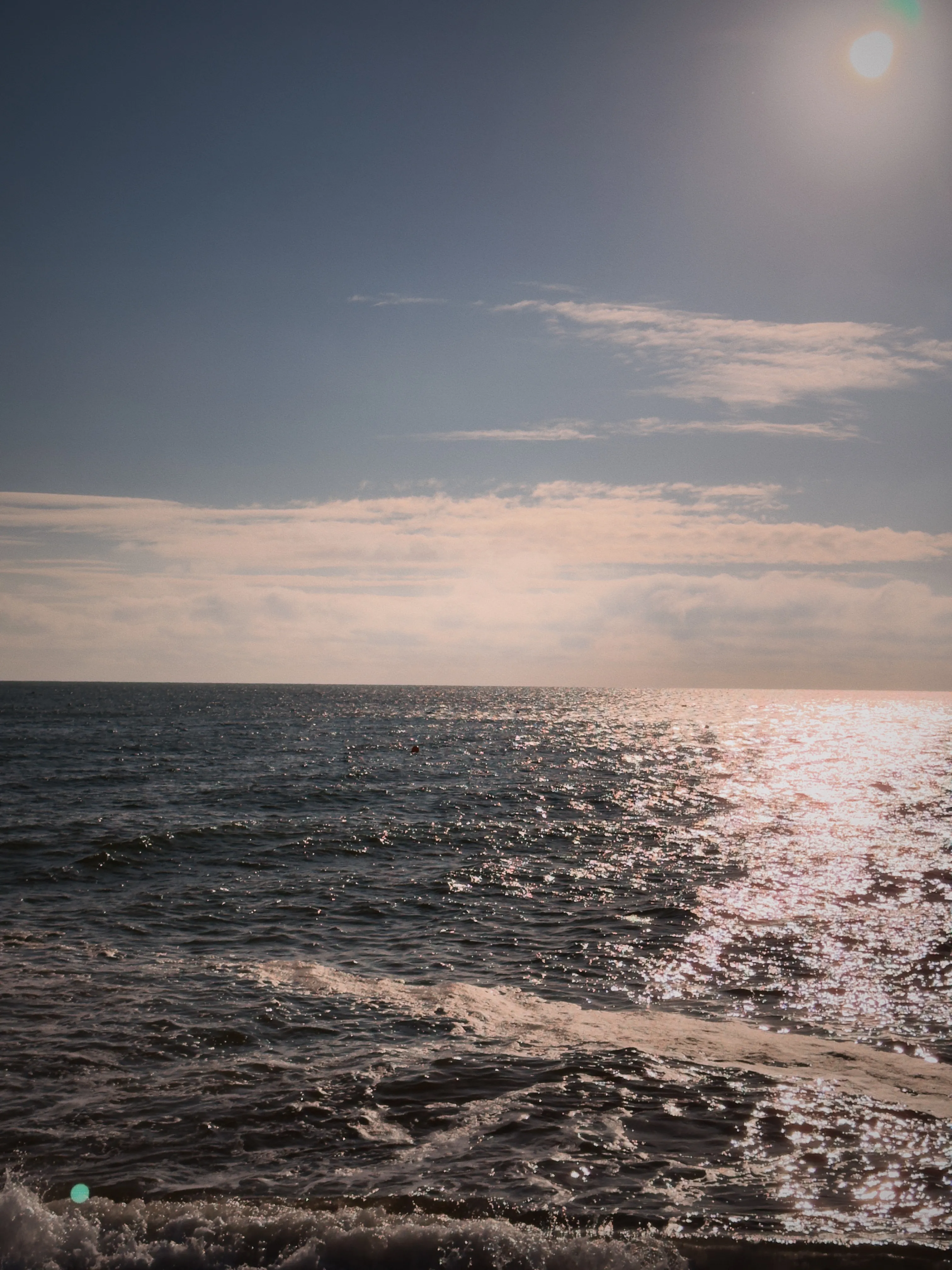 Bright autumn morning with strong lens flare, deep teal sea calm and glittering, stone pier railing at lower left