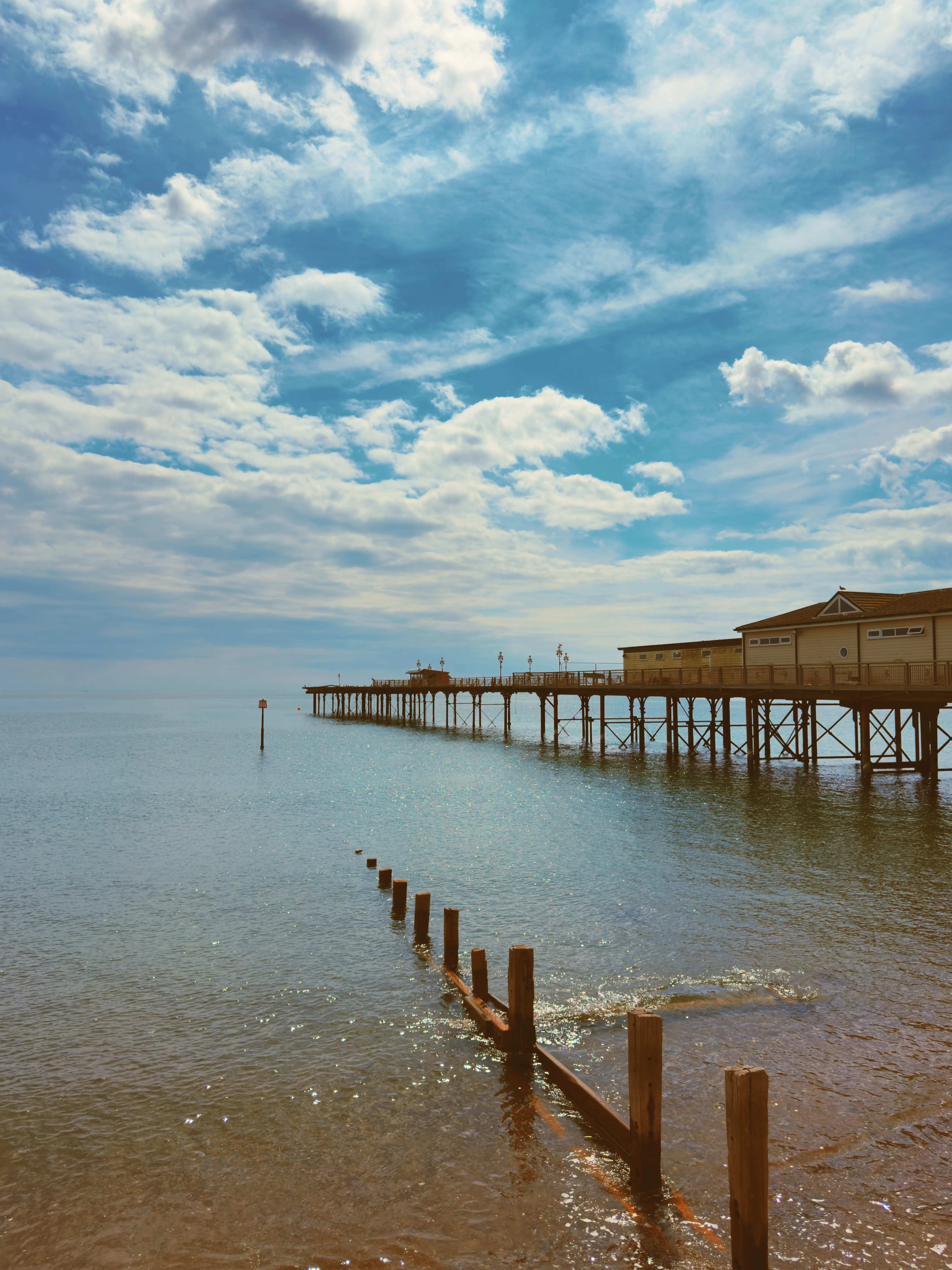 Teignmouth Pier extending left to right across the mid-frame in full summer, wooden groyne posts rising from shallow water, calm blue sea, warm summery light