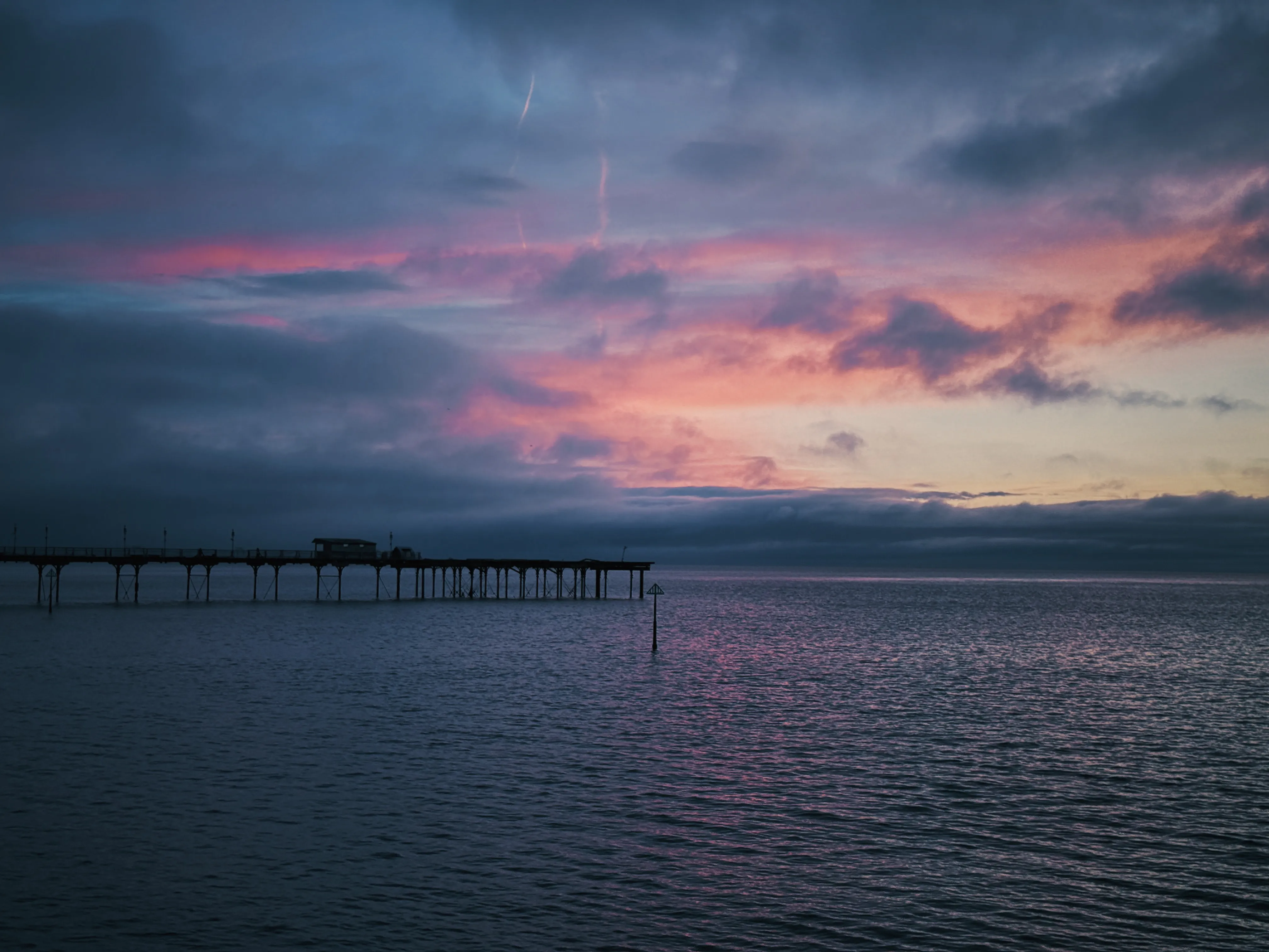 Pre-dawn, pier silhouetted on the left over glassy high water, soft pink-coral glow at the horizon