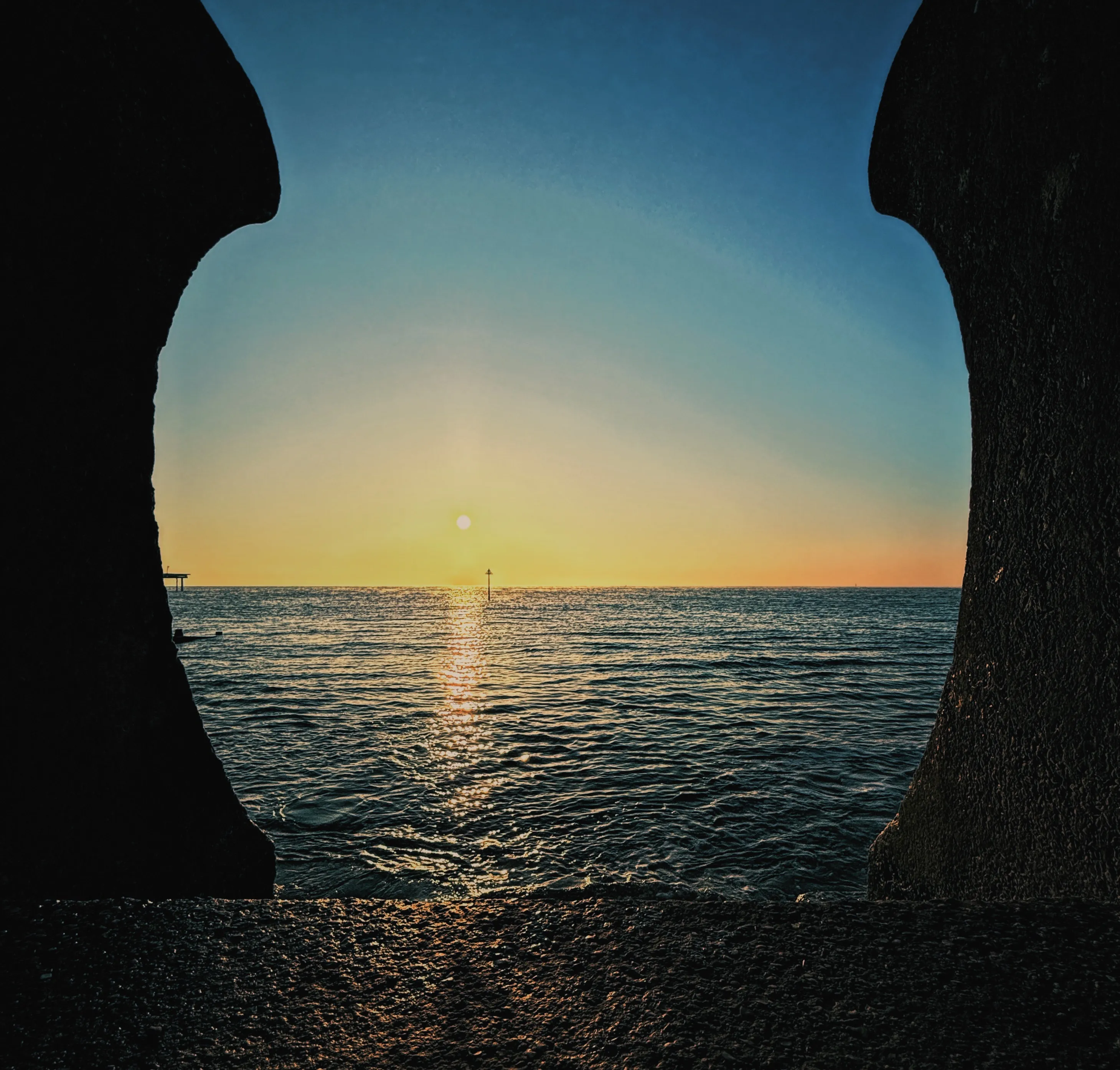 Sunrise framed through a stone arch or sea-wall gap, calm rippled water with a sun path, a distant navigation marker