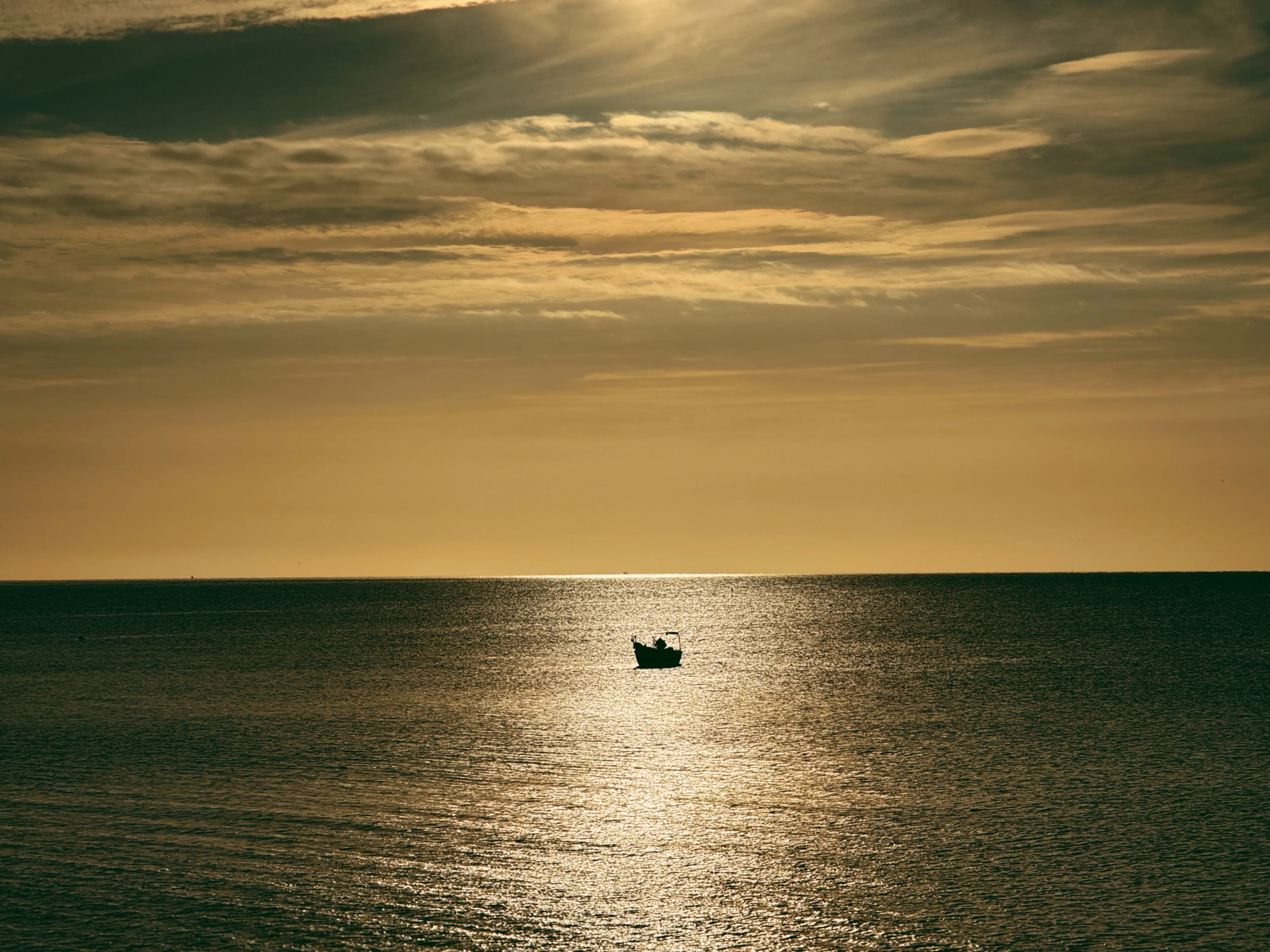Warm golden-brown morning sky, flat glassy sea with a fishing boat silhouetted in the sun's reflection